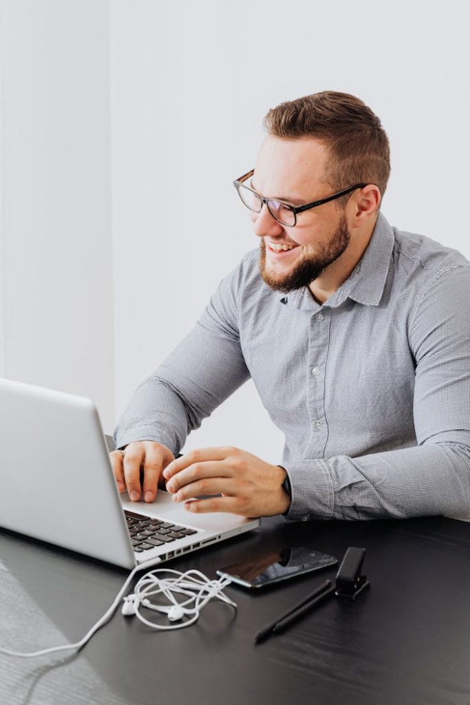 Portrait of a smiling bearded man using a laptop in a modern office setting, bright light.